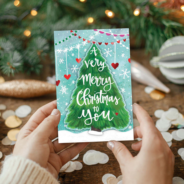 Person holding a Christmas card with a tree design and festive text, surrounded by Christmas decorations.