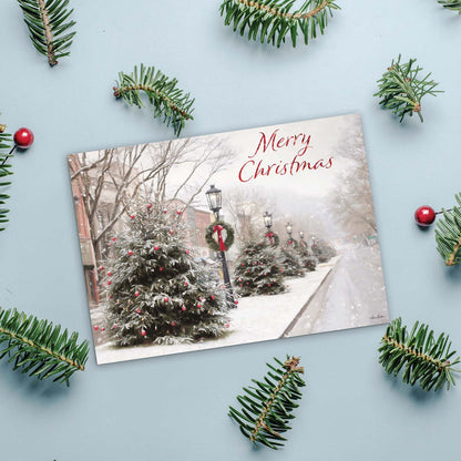 Christmas card with snowy street scene and 'Merry Christmas' text, surrounded by pine branches and berries on a light blue background.