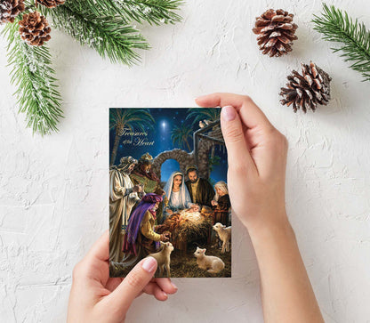 Person holding a Christmas card with a nativity scene design, surrounded by pine branches and pinecones on a light background.